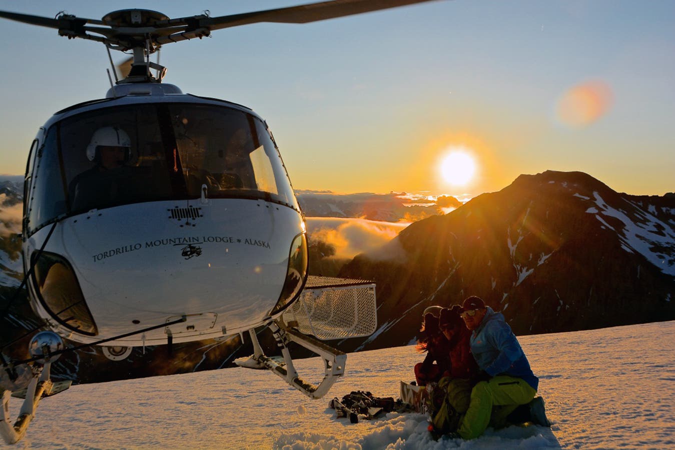 A group of skiers watches as the heli takes off under the midnight sun.