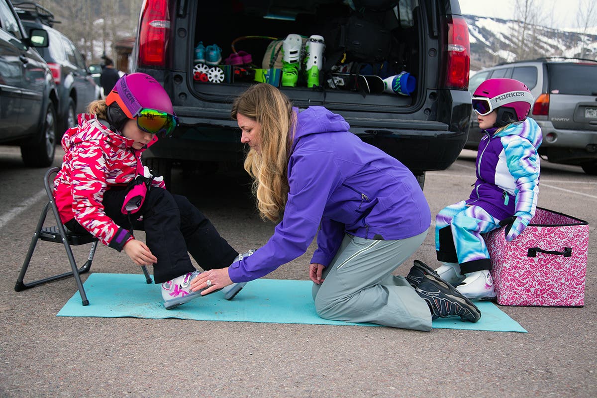 ExtrasForCar Mother helping two daughters put on ski gear in the parking lot of a ski resort.