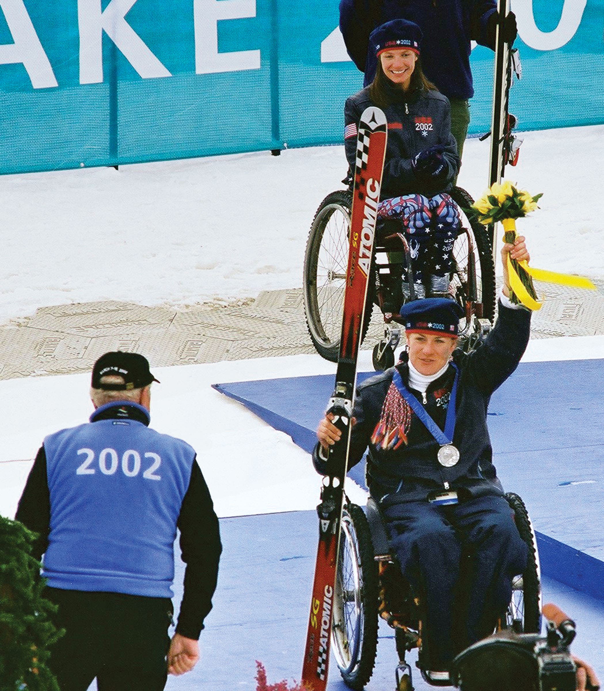 SKI1119-PRO4-MuffyDavis_1 copy Muffy Davis sitting in her wheelchair at the 2002 Salt Lake Paralympics holding a bouquet and a pair of red atomic skis with a silver medal around…