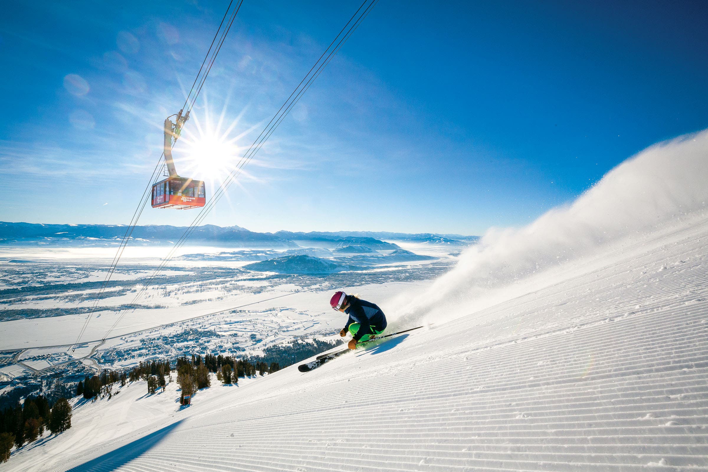 Skier skiing down well groomed slope with a gondola above the skier that says Jackson Hole