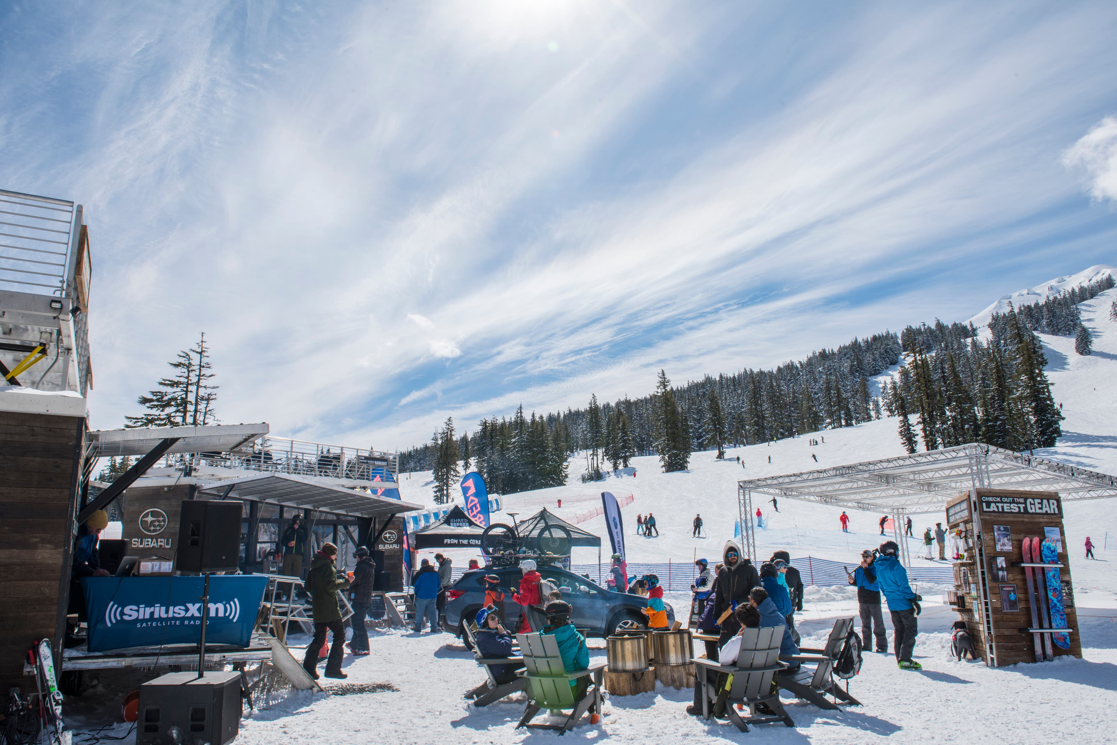 People sit in adirondack chairs at the base of Mt Bachelor on a sunny day. Stages and event tents surround them.