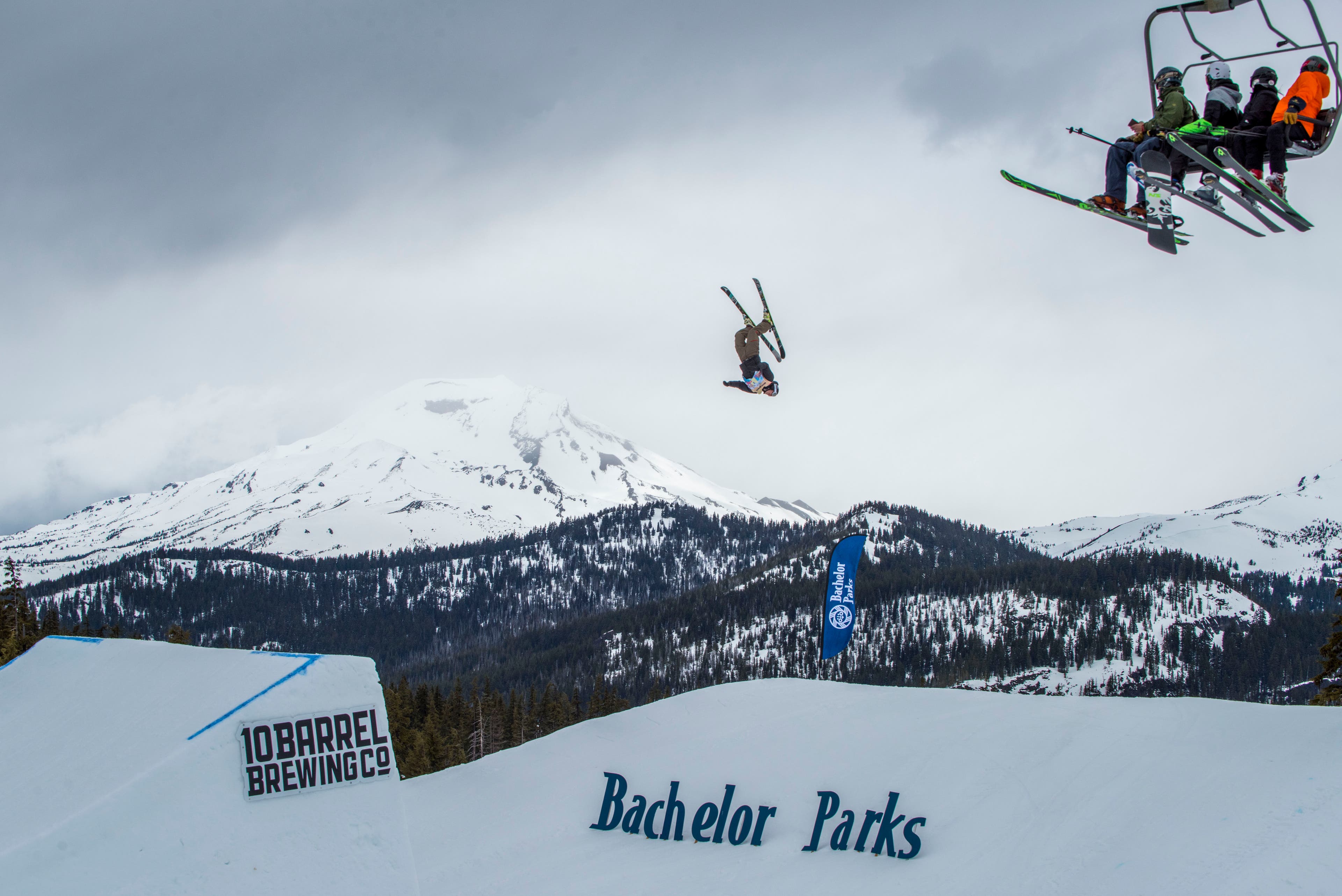 Four people sit on a chairlift and watch a person flipping off a ski jump.