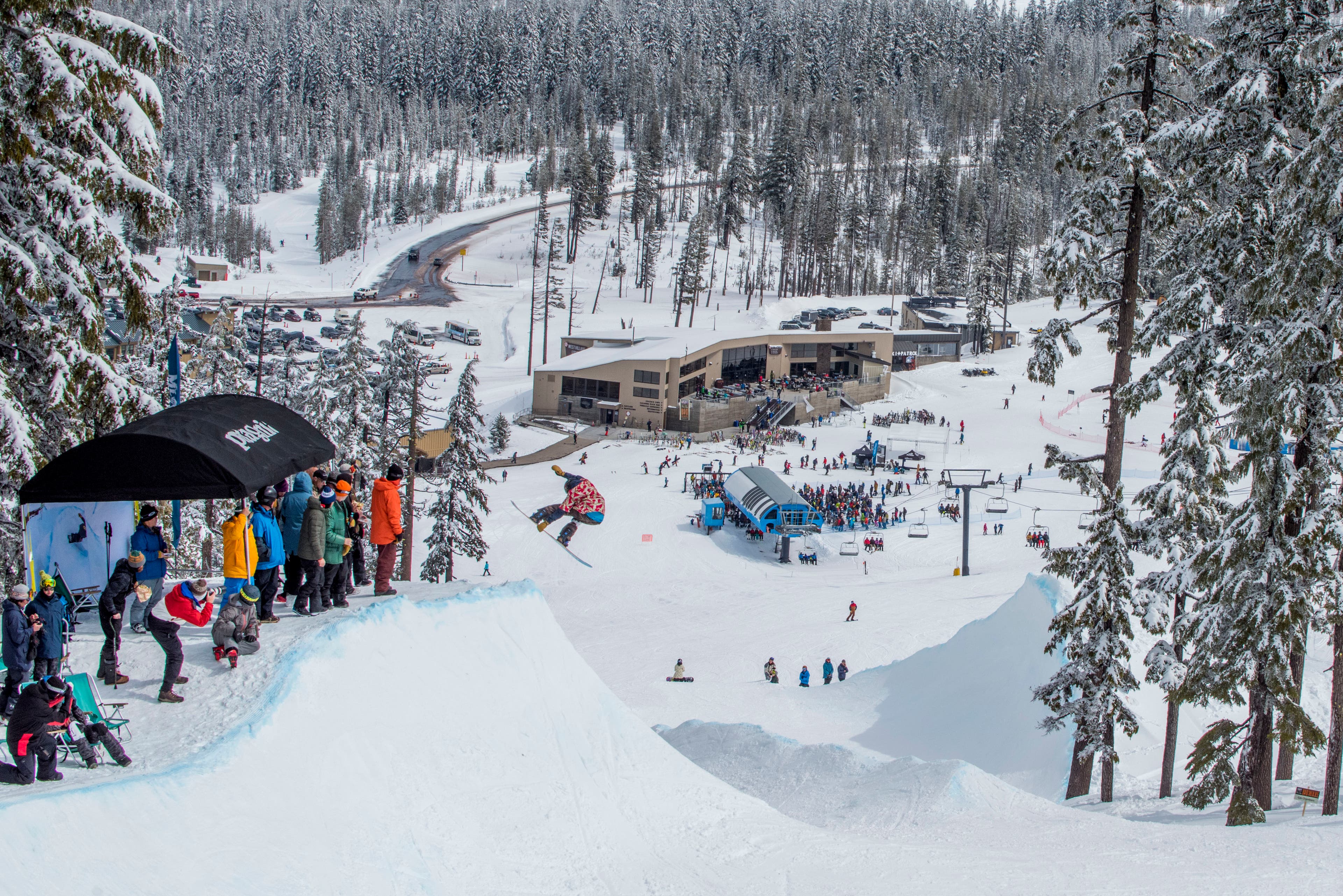 Snowboarder in red jacket grabs their board at the top of a pipe while a crowd of people watches.