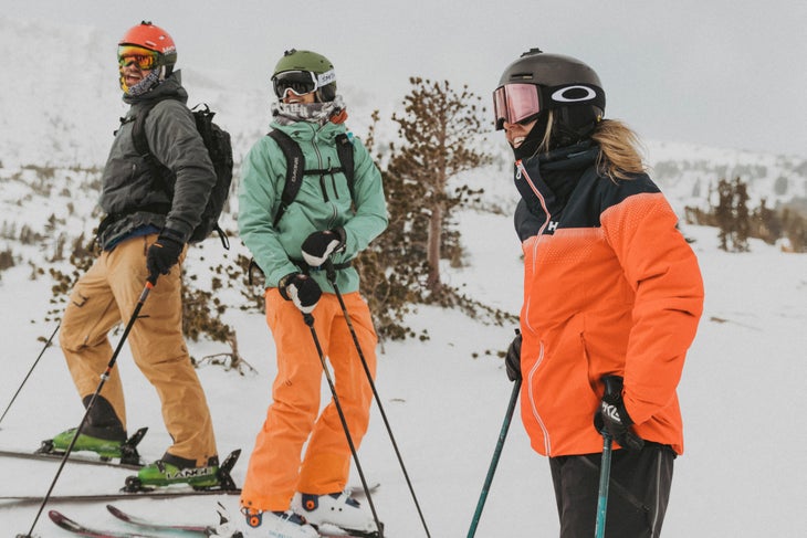 CrewVibes_MammothMountain Three people stand on skis at Mammoth Mountain and laugh. All wearing different combinations of black, orange, and light teal. Rolling hills covered…