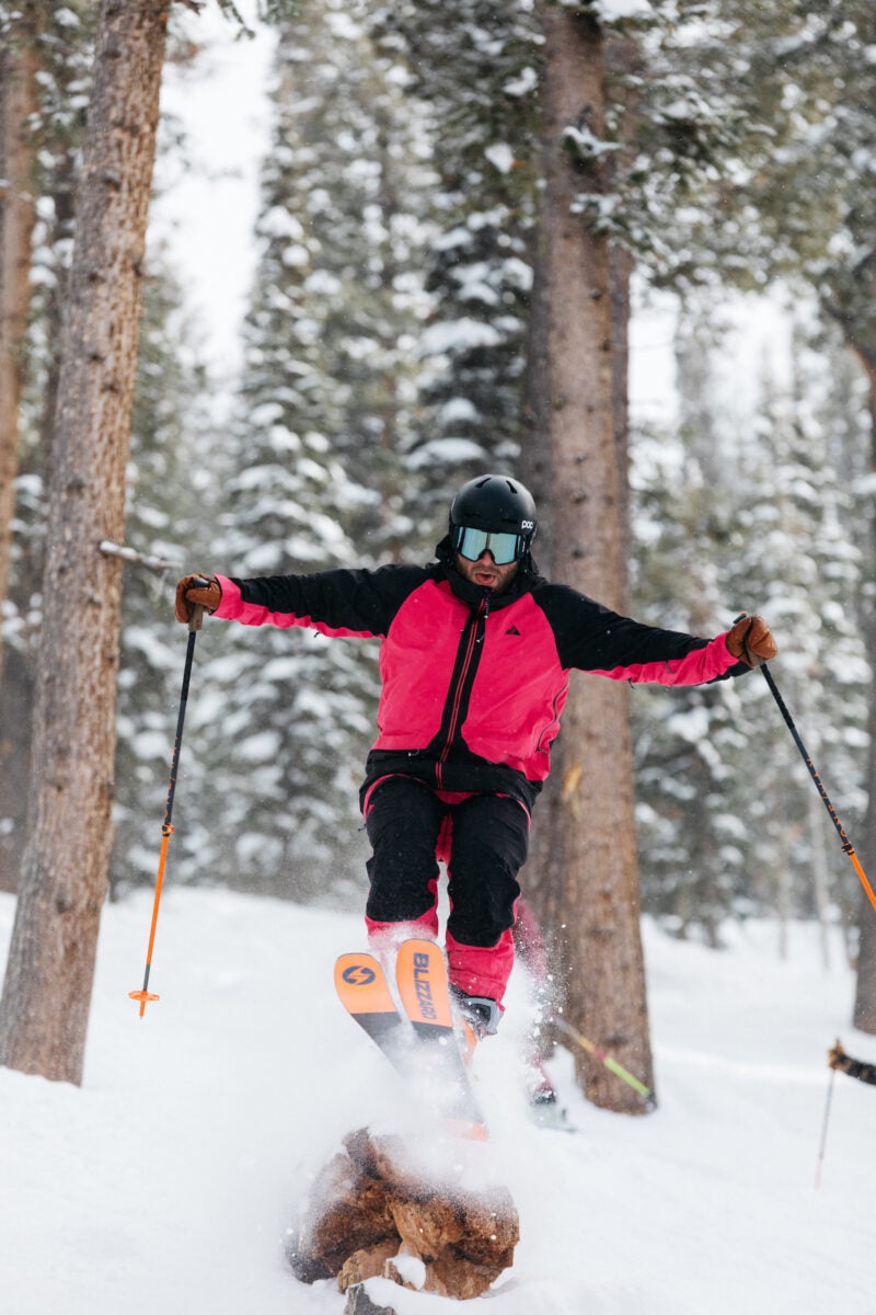 A skier in pink and black jumps off a downed log in the trees in Sun Valley, Idaho