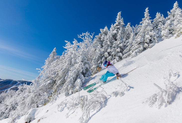 Skier in blue pants and white jacket rips down mountain on a bluebird day.