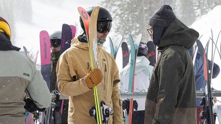 Skier holding a pair of skis talking to another skier in front of ski racks