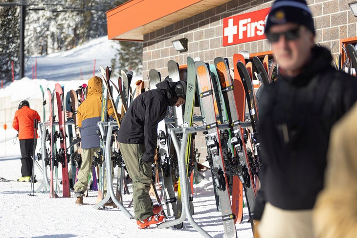 Skis on racks on snow with skiers getting ready