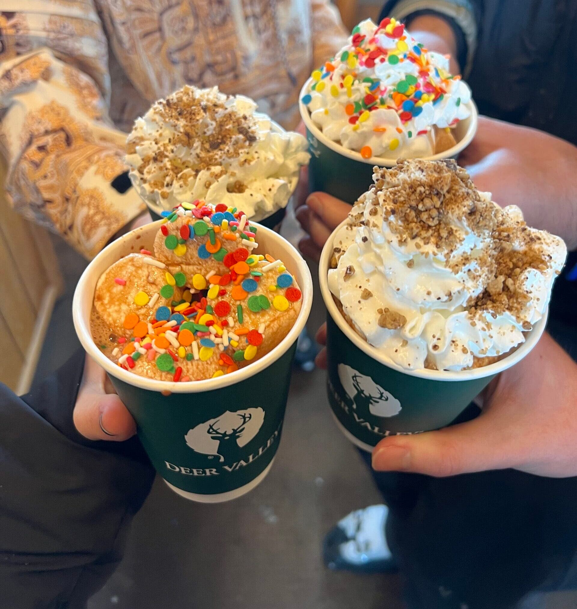 four hot chocolate cups with marshmallows, whip cream, and sprinkles on top.