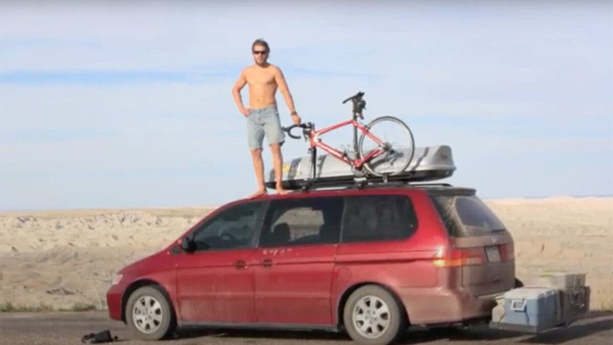Man posing on top of red minivan with road bike and roof box on top
