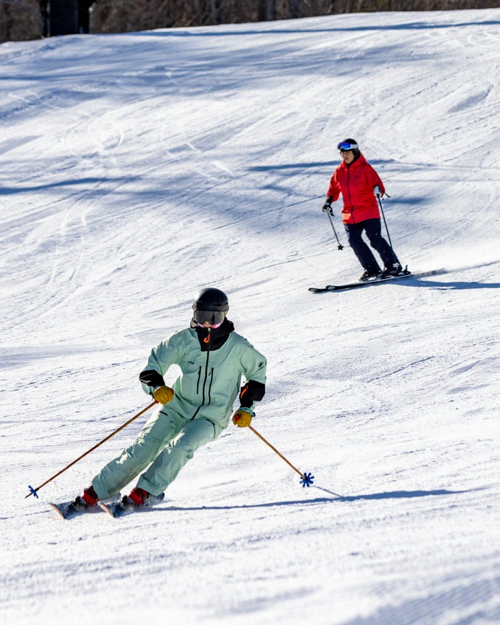 Two skiers, the front one in a mint green kit and the following in red ski down the mountain