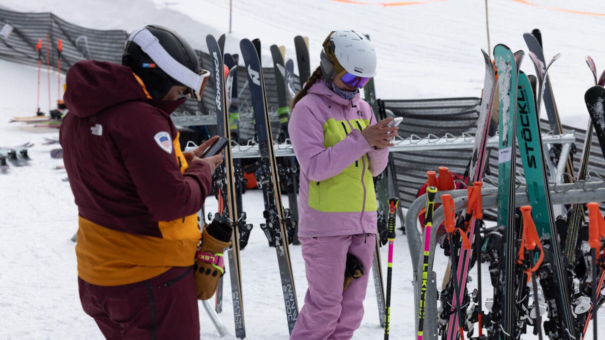 Two skiers typing on phones by ski racks at ski resort base