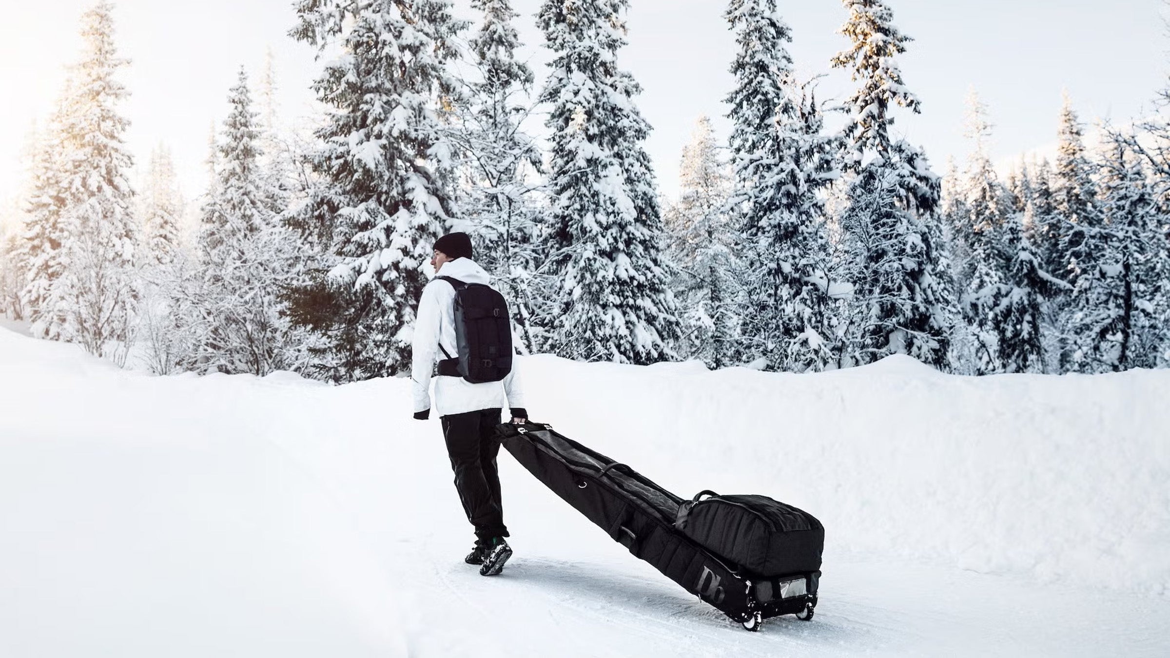 Man in white jacket black pants pulling black roller ski bag and boot bag along snowy road