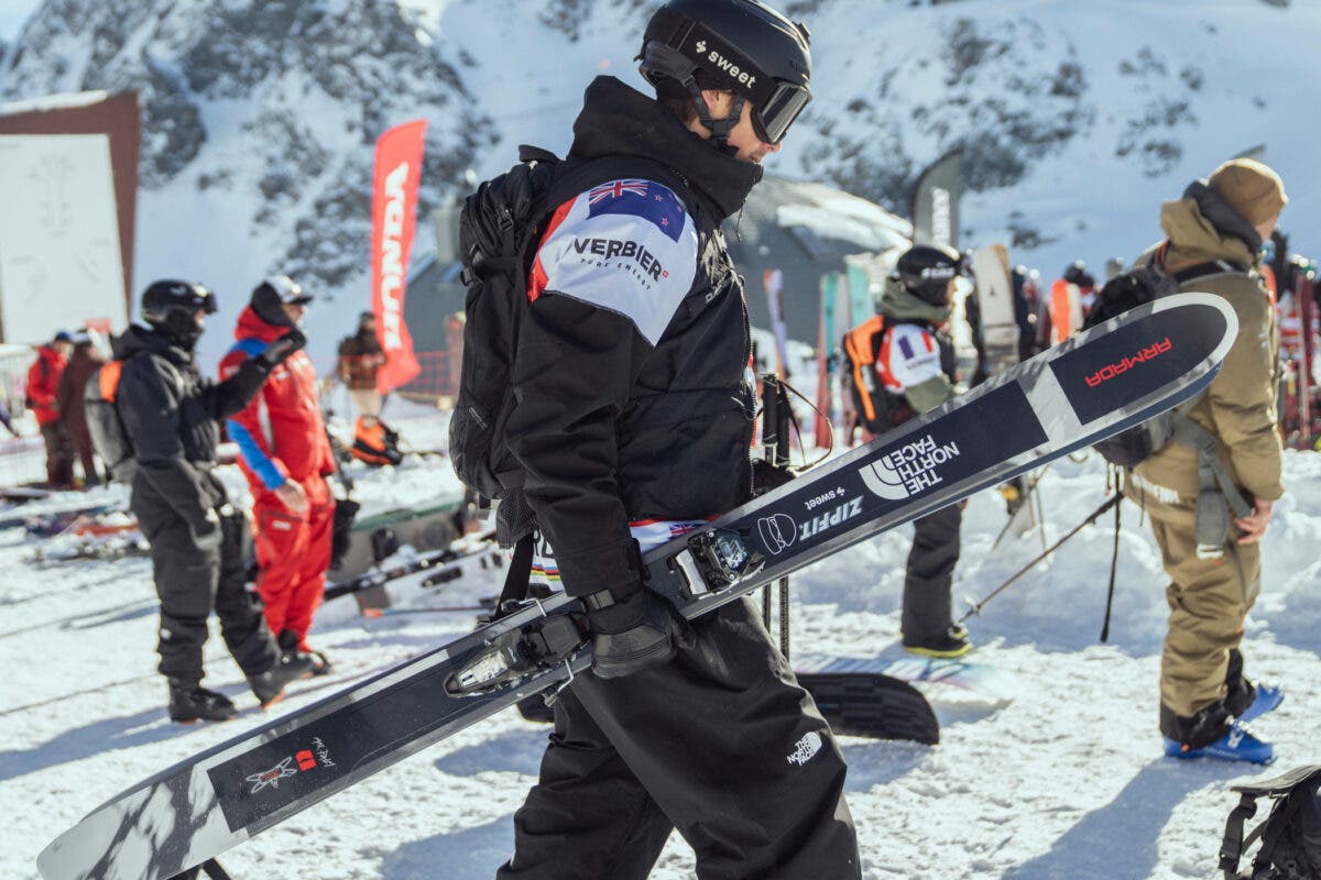 Skier wearing a New Zealand flag patch and Verbier competition bib carries a pair of Armada Antimatter skis at a Freeride World Tour event.
