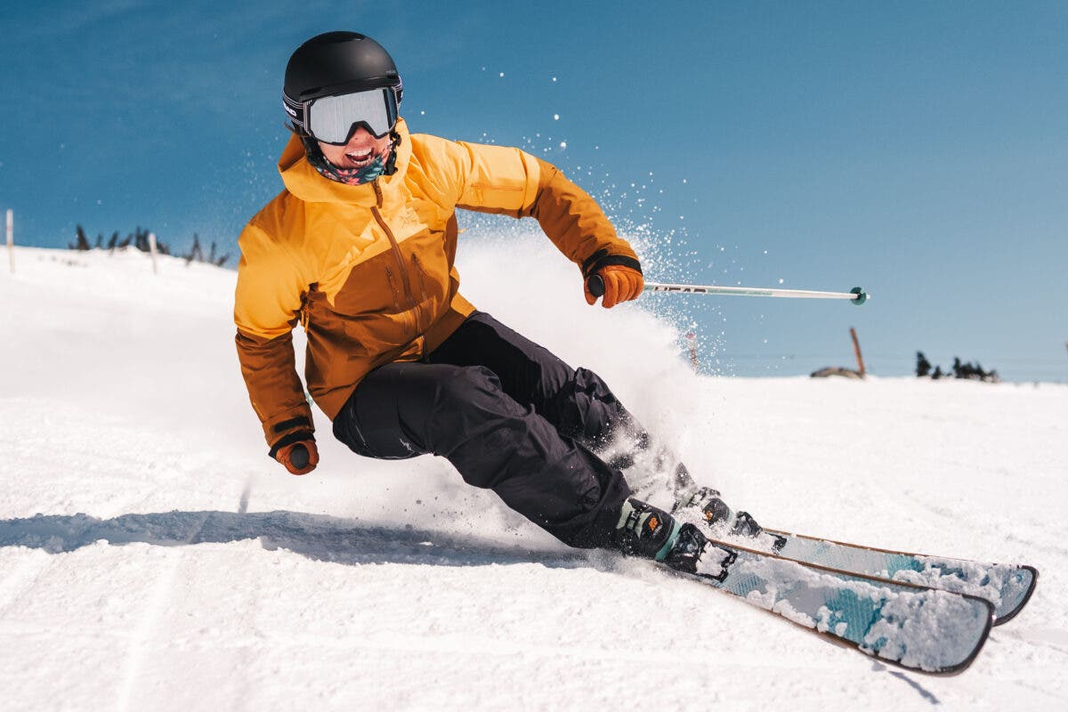 Woman in yellow and orange jacket carving down ski slope on Head skis on sunny spring day