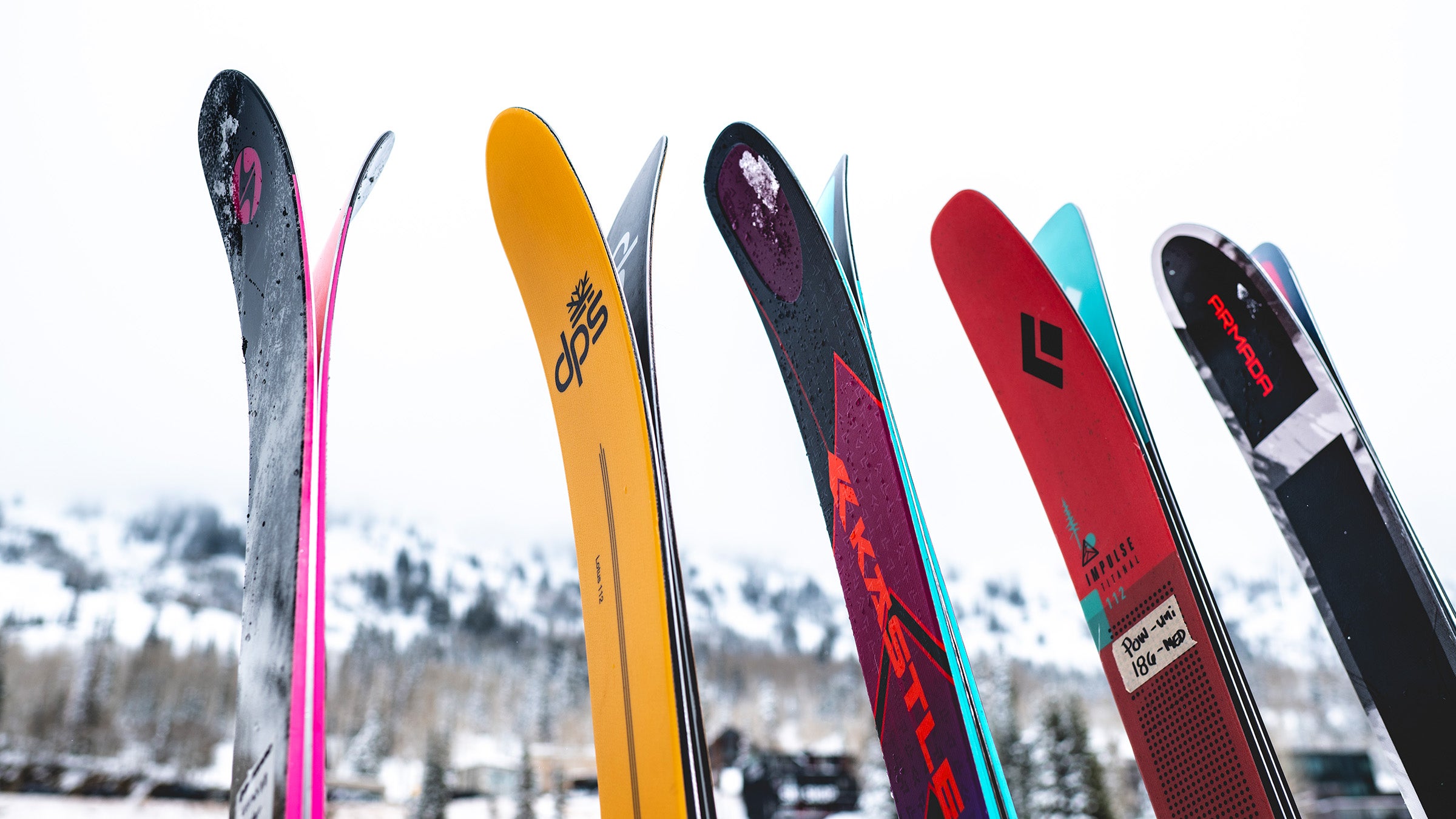 A row of skis with visible tip rocker profiles standing upright against a snowy mountain backdrop, including models from DPS, Kastle, Black Diamond, and Armada.